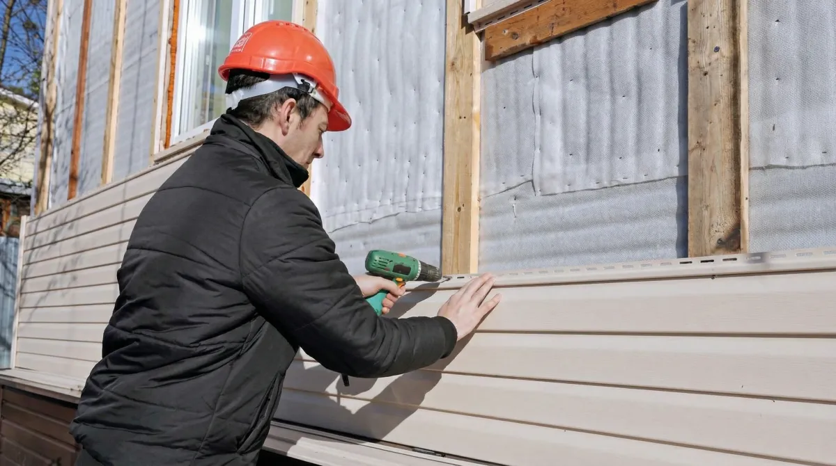 Siding replacement professional using a power drill to install new tan vinyl siding on a home exterior in Portland, OR